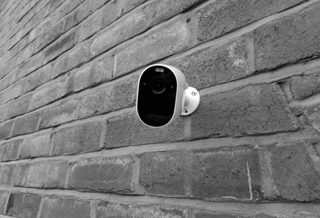 A black and white photo of a security camera mounted on a brick wall.